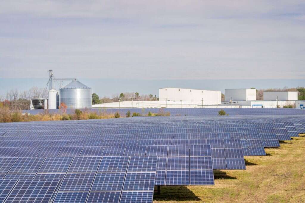 Large solar panel field with warehouse buildings in the background under a clear sky