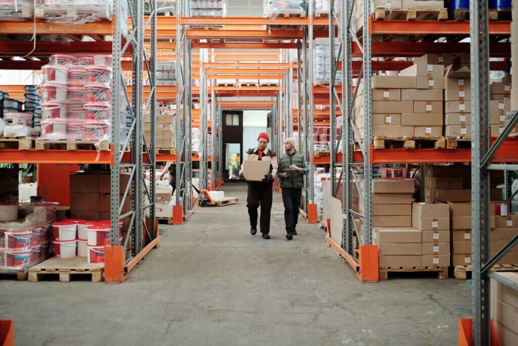 Two warehouse workers handling a package beside shelves stocked with boxed products